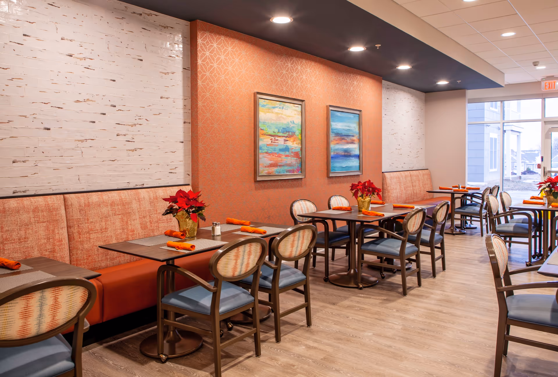 Bright dining room with multiple tables and chairs, banquette seating, colorful wall art, and poinsettia centerpieces.