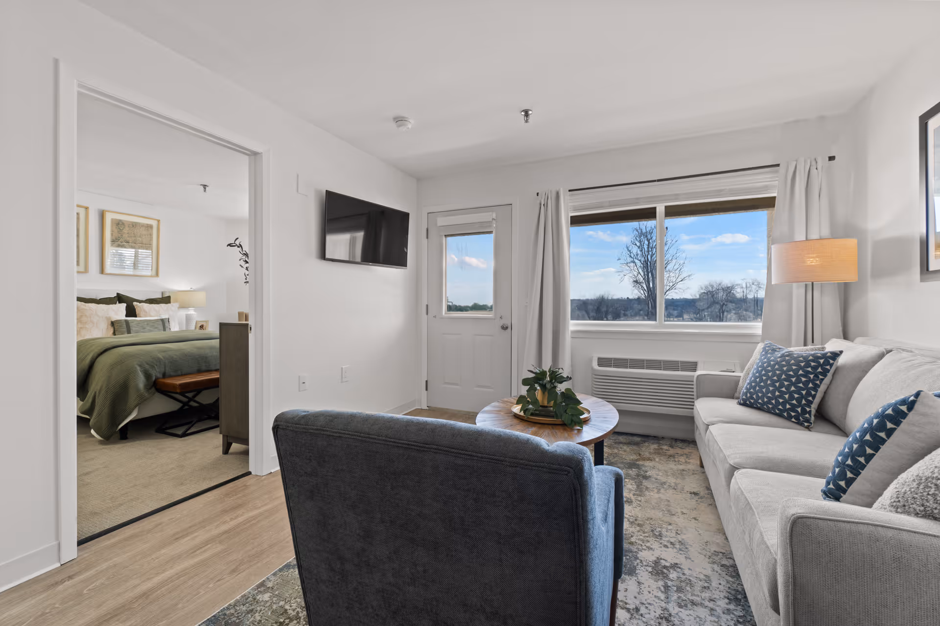 Bright living room with a sofa, armchair, round coffee table, wall-mounted TV and a window with an outdoor view; a bedroom is visible through an open doorway.