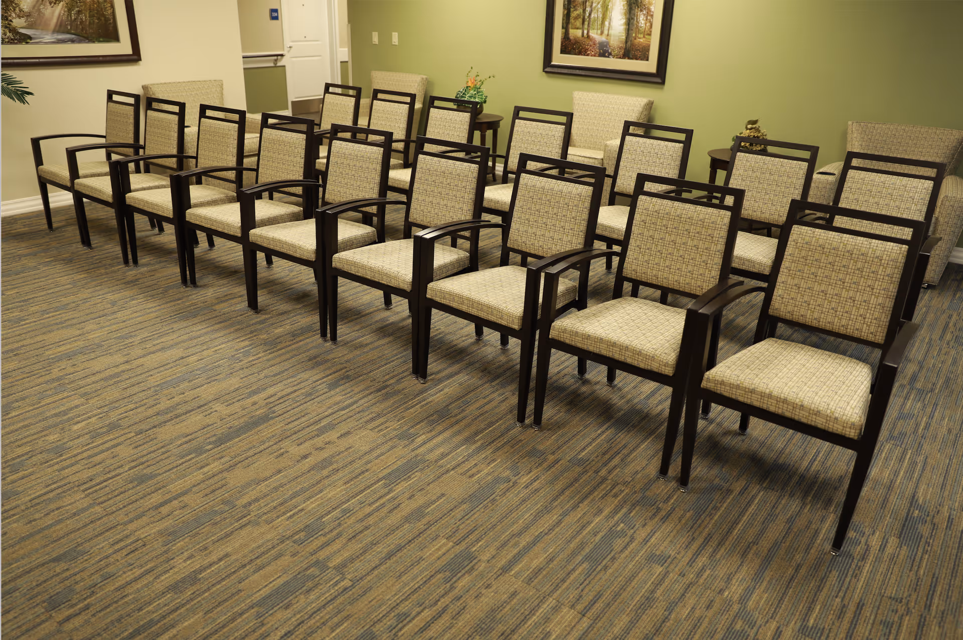 Rows of upholstered wooden chairs arranged in a meeting or waiting room with green walls and framed artwork.