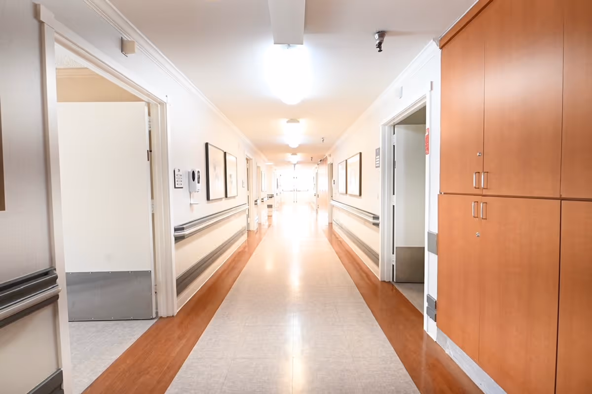 A long, well-lit hallway in a healthcare facility with light-colored walls, wooden cabinets on the right, handrails along both sides, and several open doors leading to rooms.