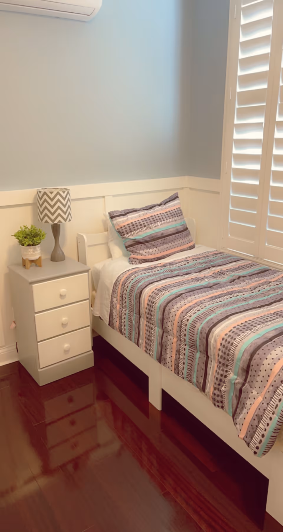 A neatly made single bed with a striped and patterned bedspread and matching pillow in pastel colors. Next to the bed is a small white nightstand with three drawers, a gray and white chevron-patterned lamp, and a small green potted plant. The room has light blue walls, white wainscoting, wooden flooring, and white plantation shutters on the window.