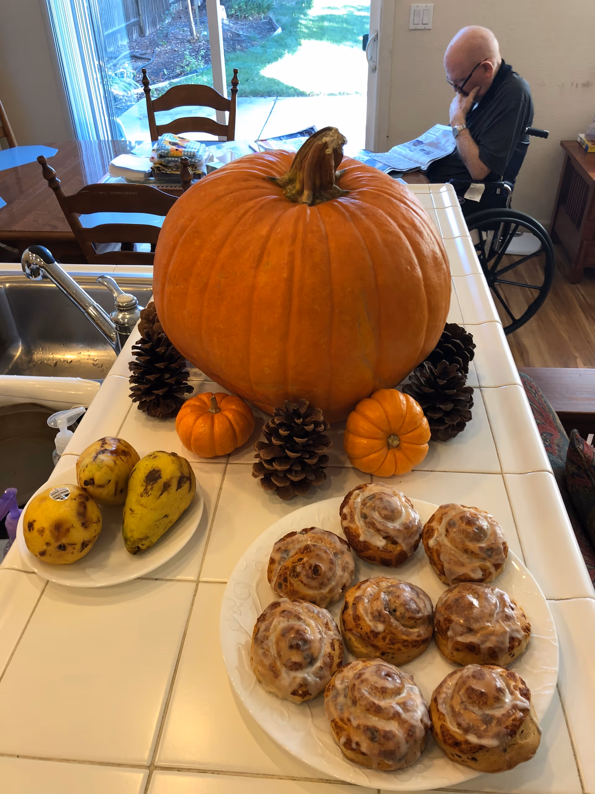 A kitchen island topped with a large pumpkin, small gourds, pine cones and a plate of iced cinnamon rolls, with a man in a wheelchair reading in the background.