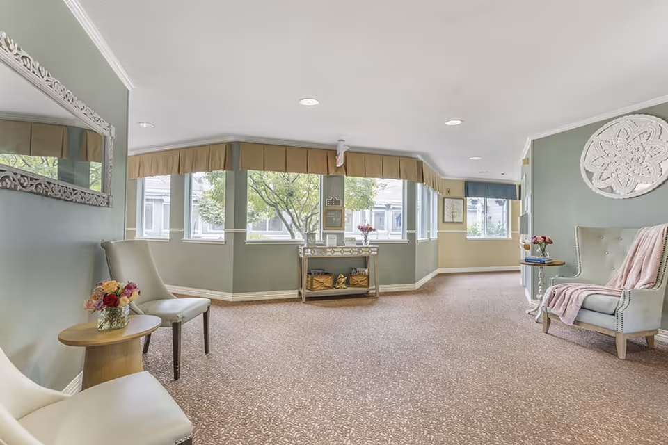 A bright and spacious interior common area with large windows letting in natural light. The room features light green walls, beige carpet, and several chairs with small tables adorned with flower arrangements. Decorative wall art and a large mirror add to the cozy ambiance.