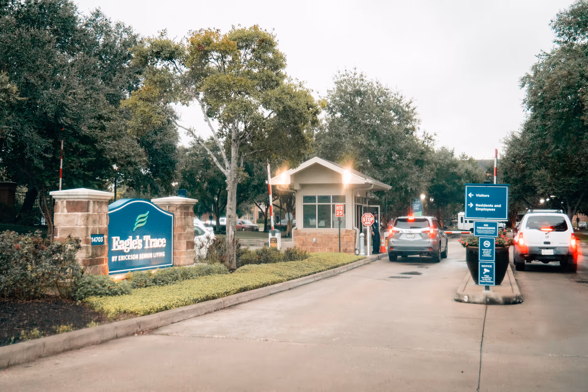 Entrance to Eagle's Trace senior living facility with a guardhouse, two cars waiting at the gate, and signage directing visitors and residents. The area is surrounded by trees and landscaped greenery.