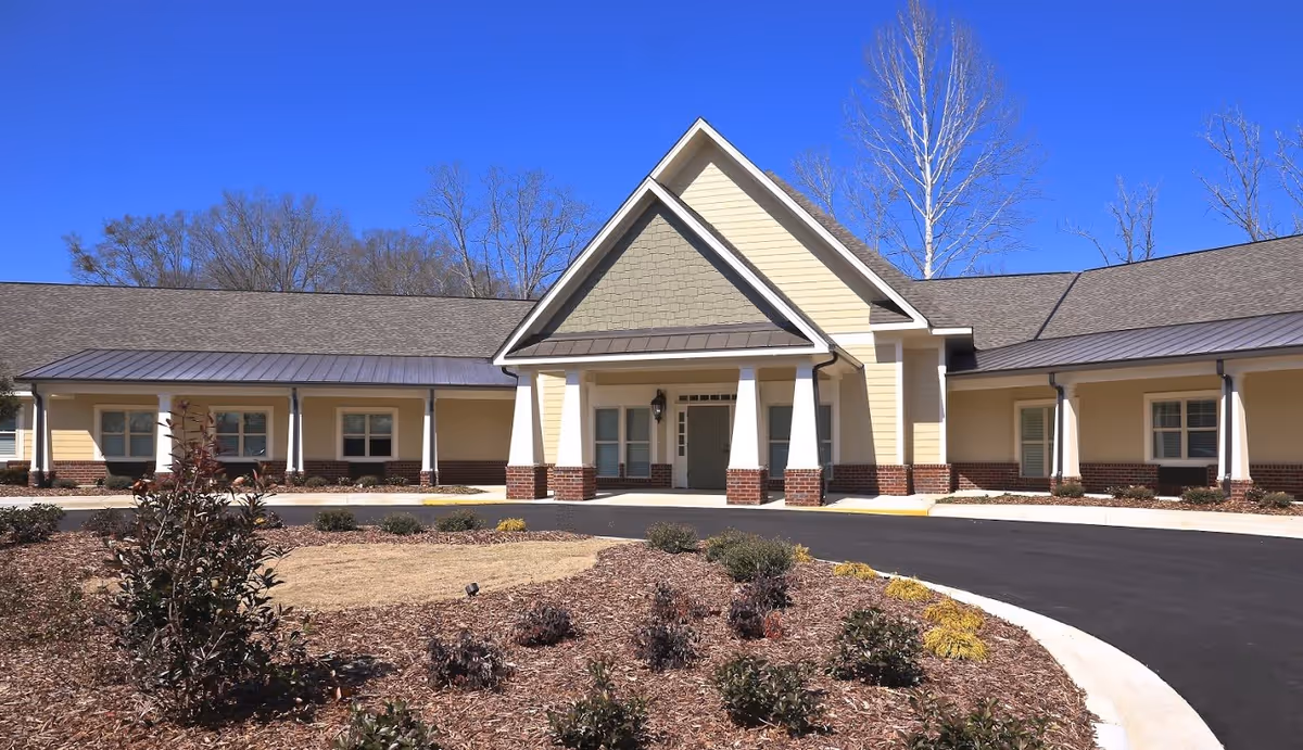 Front exterior view of a single-story building with a peaked roof and a covered entrance supported by white columns. The building is surrounded by landscaped grounds with mulch and small shrubs under a clear blue sky.