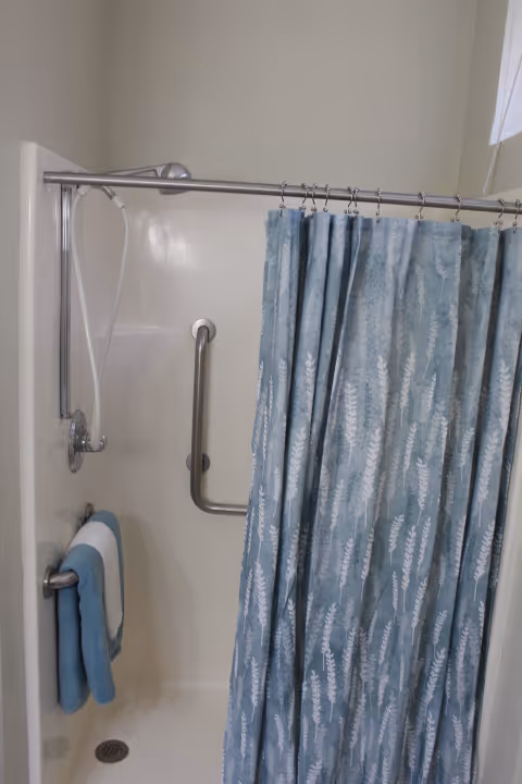 A shower area with a silver showerhead, a grab bar on the wall, and a blue shower curtain with a white leaf pattern. There are two towels, one blue and one white, hanging on a towel bar inside the shower.