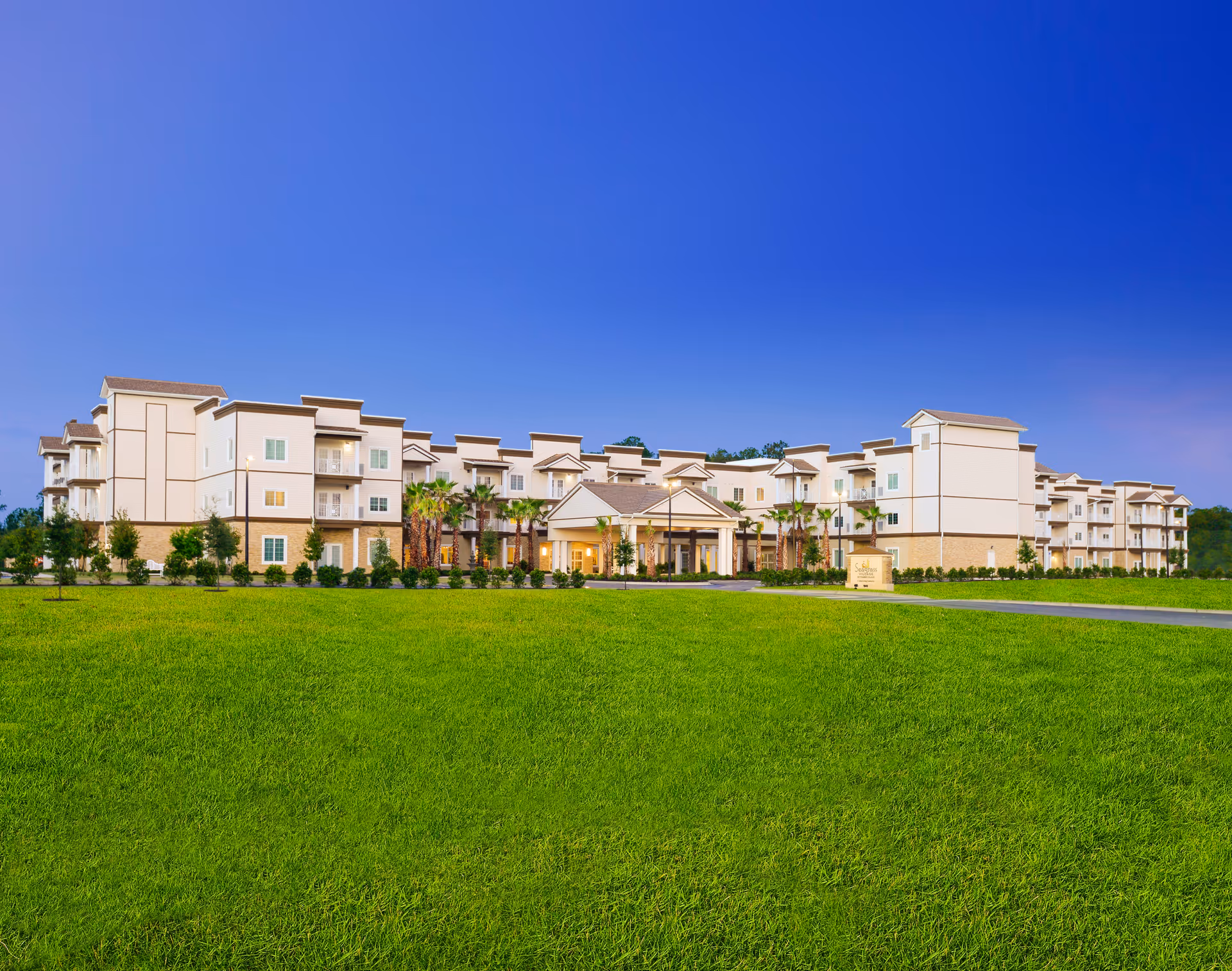 Wide exterior view of Seagrass Village of Fleming Island, a large multi-story senior living facility with beige and white walls, multiple balconies, palm trees, and a well-maintained green lawn under a clear blue sky.