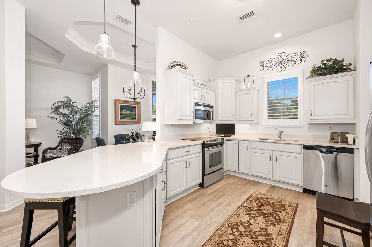 Bright and modern kitchen with white cabinetry, stainless steel appliances including a stove, microwave, dishwasher, and refrigerator. The kitchen features a large white countertop island with two pendant lights hanging above it. There is a window with white shutters above the sink, a decorative wall piece above the window, and a patterned rug on the wooden floor. In the background, part of a dining area with a chandelier, a framed painting, and a potted plant is visible.