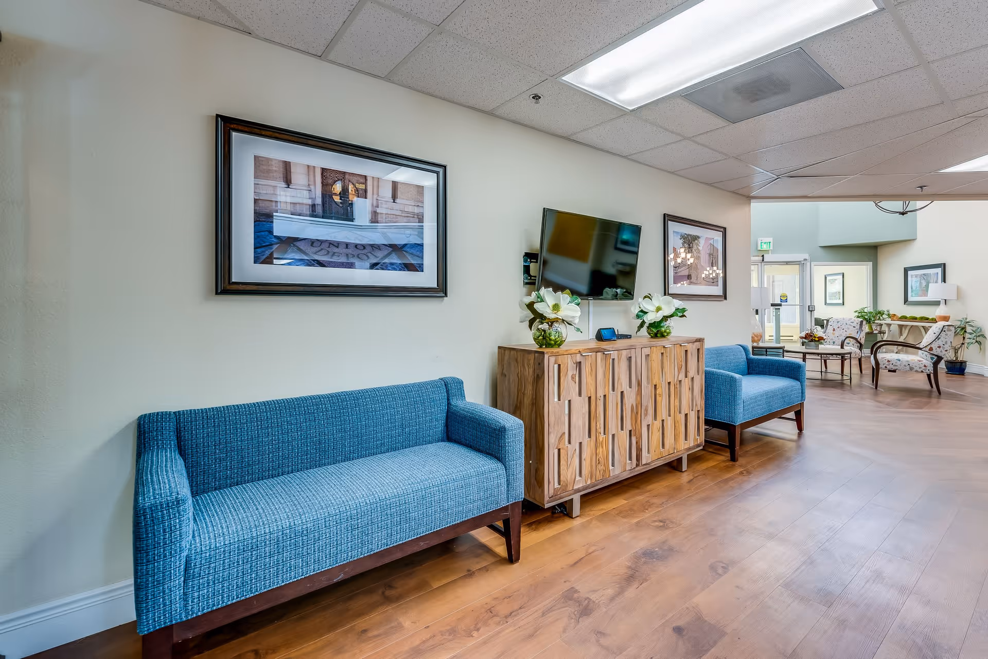 A senior living facility common area with two blue upholstered sofas, a wooden cabinet with decorative flowers, a wall-mounted flat screen TV, framed artwork on the walls, and additional seating with armchairs and a coffee table in the background.