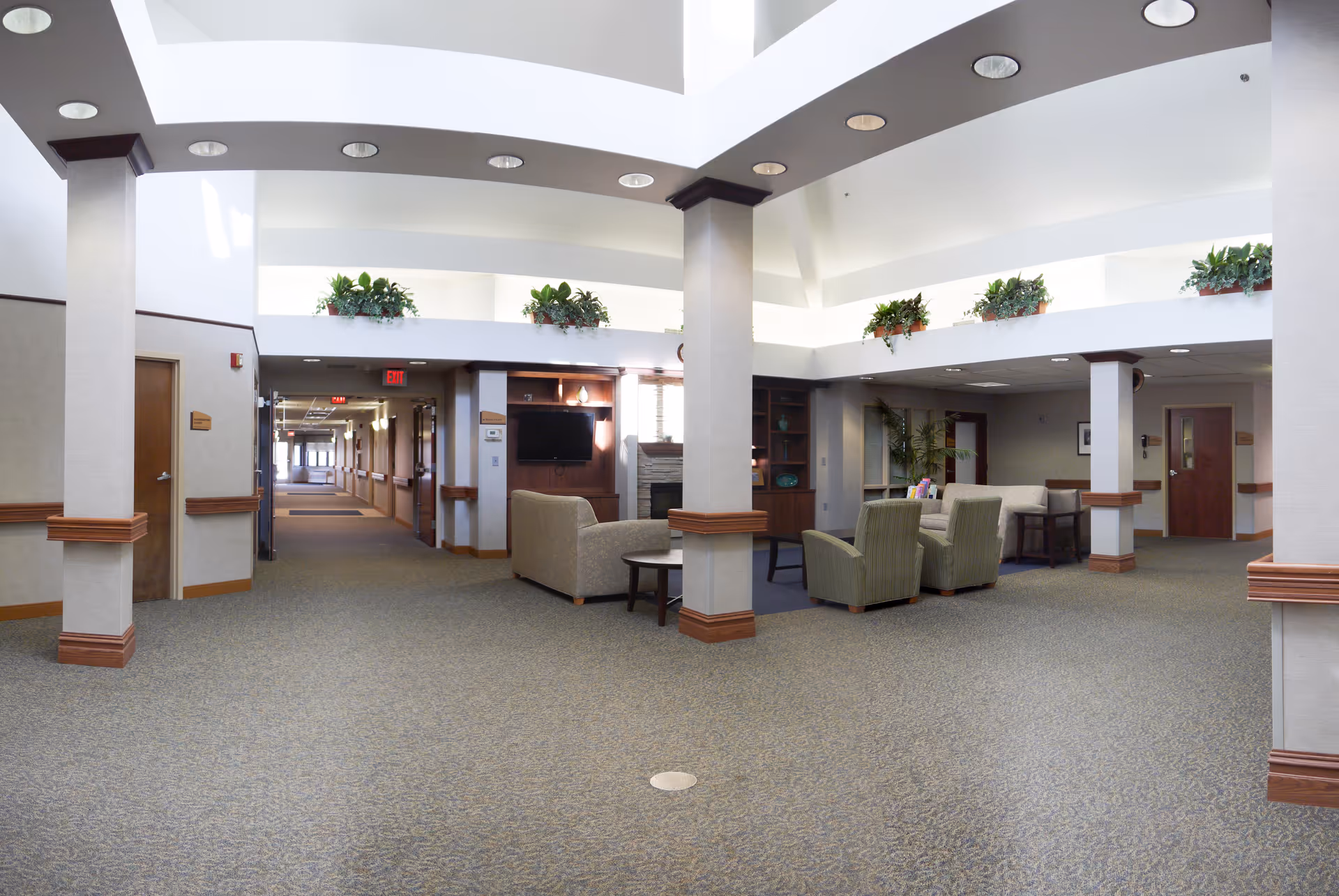 A spacious and well-lit common area in a senior living facility with beige carpet, white walls, and wooden trim. The area features comfortable seating including armchairs and sofas arranged around small tables. There are large columns, built-in shelves with decorative items, and potted plants on ledges near the ceiling. A hallway extends from the common area with doors along the sides and exit signs visible.