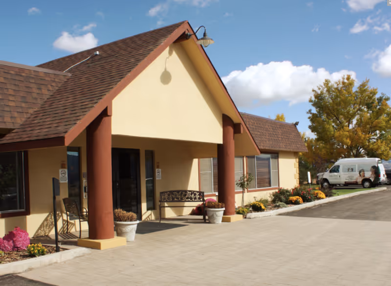 Exterior view of Monument Health Richfield facility showing the entrance with a covered porch supported by two large columns, a bench, potted plants, and a parking area with a white van. Trees with autumn foliage are visible in the background under a partly cloudy sky.