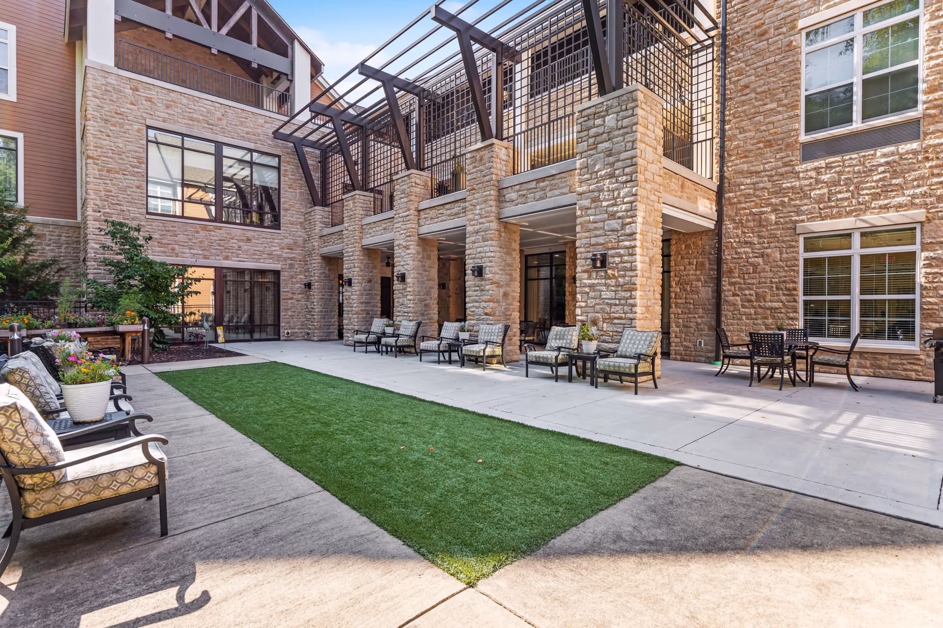 Outdoor courtyard area of a senior living facility with stone walls, large windows, and a covered patio supported by stone pillars. The courtyard features a strip of artificial grass in the center, surrounded by concrete walkways. Several cushioned chairs and small tables are arranged along the sides, with potted plants and flowers adding greenery to the space.