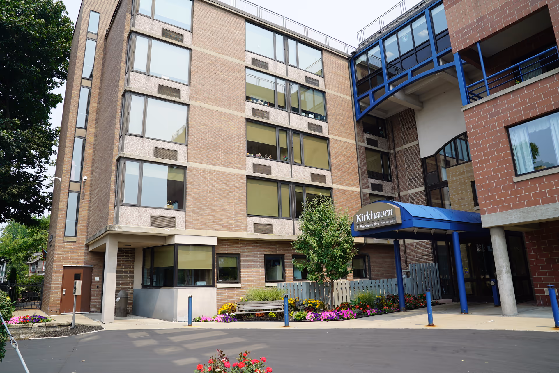 Exterior view of a multi-story brick building with large windows and a blue awning entrance labeled 'Kirkhaven'. There are flower beds and a small tree near the entrance, with a paved driveway in front.