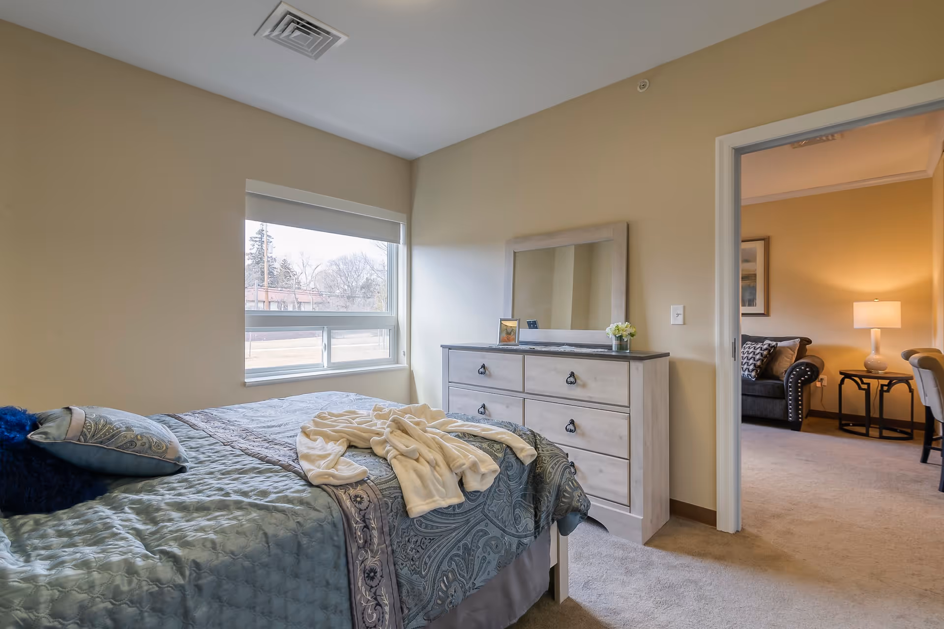 A bedroom with a neatly made bed covered in a blue patterned comforter and pillows, a light-colored dresser with a mirror on top, and a window letting in natural light. Through an open door, a living room area with a sofa, side table, lamp, and chairs is visible.
