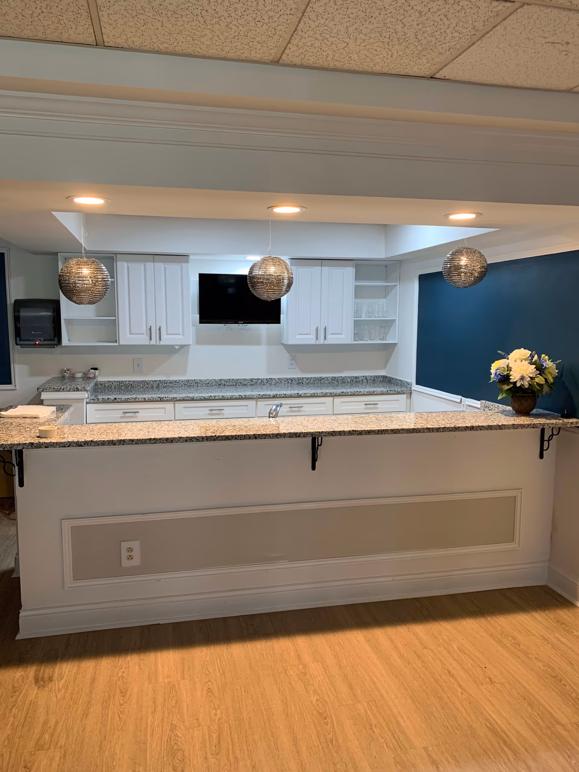Interior view of a kitchen area with a granite countertop bar, white cabinets, a mounted flat-screen TV, three spherical pendant lights hanging from the ceiling, and a vase with white and blue flowers on the right side of the counter. The floor is wooden, and the walls are painted white and dark blue.