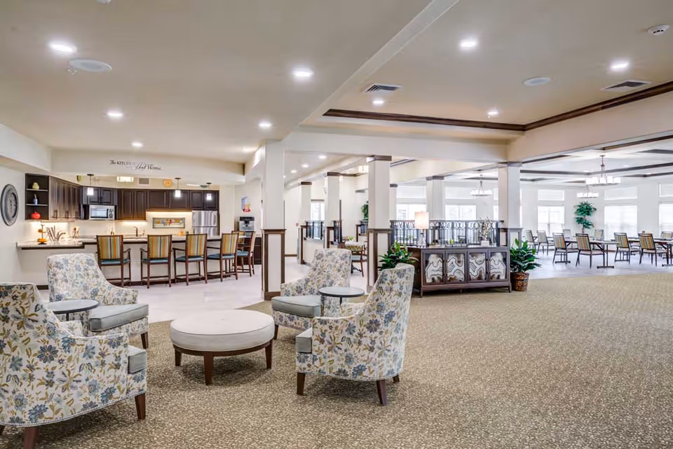 Open, airy senior living common area with floral upholstered chairs around a round ottoman, a kitchen counter with barstools, and a dining area in the background.