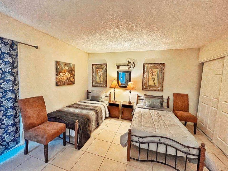 Bright twin bedroom with two single beds separated by a nightstand holding two lamps, framed artwork on the walls, and two chairs.