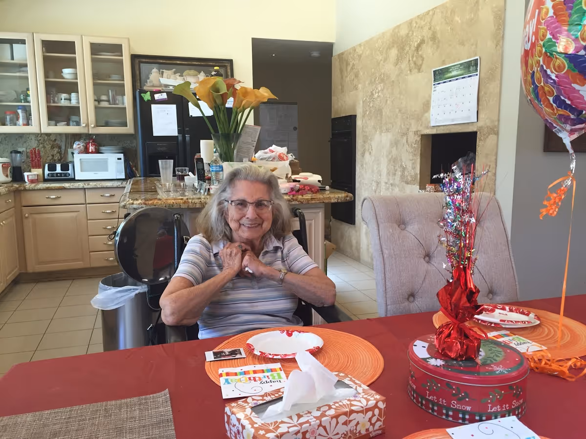 An elderly woman sitting in a wheelchair at a dining table in a kitchen area. The table is covered with a red tablecloth and has birthday decorations, including a balloon, a gift box, and a birthday card. The kitchen in the background has light-colored cabinets, a microwave, a toaster, and a vase with yellow flowers on the counter.