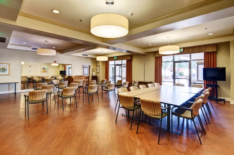 A spacious room with wooden flooring featuring multiple tables and chairs arranged for group seating. The room has large windows with brown curtains allowing natural light to enter. There are modern ceiling lights and a flat-screen TV on a stand near the windows. The room appears clean and well-lit, suitable for meetings or dining.