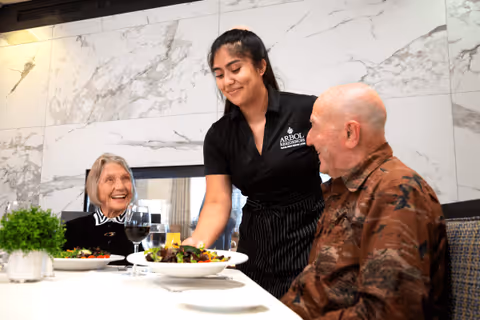 A smiling female server wearing a black Arbol Residences of Santa Rosa shirt serves plates of salad to an elderly man and woman seated at a dining table in a bright, modern dining room with marble walls.
