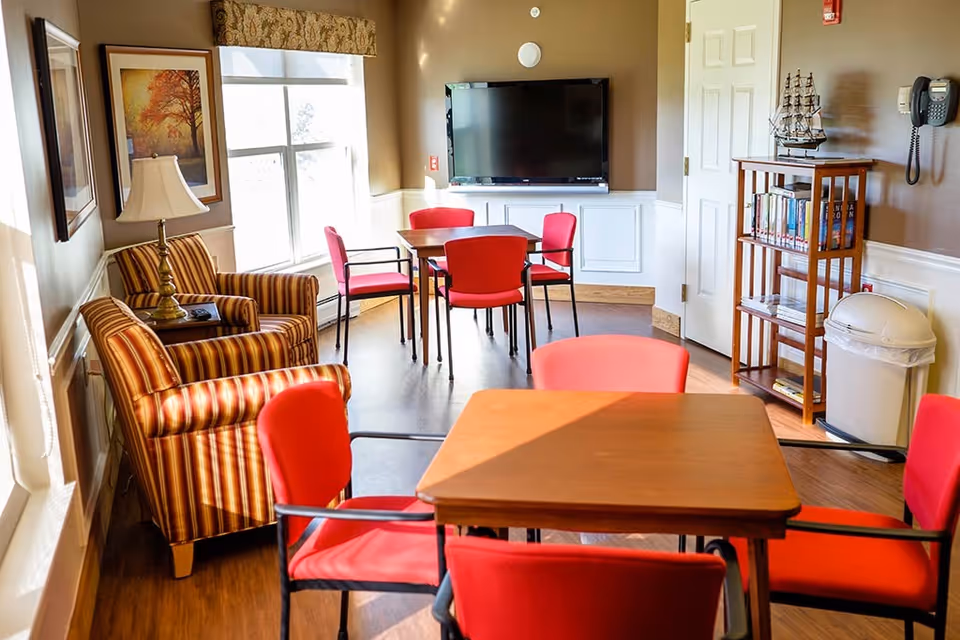 A bright common area with wooden floors featuring two square wooden tables surrounded by red cushioned chairs. There are two striped armchairs with a lamp and framed artwork on the wall near a large window. A flat-screen TV is mounted on the wall above white paneling. A small wooden bookshelf with books and a model ship sits next to a white trash can and a wall-mounted telephone.