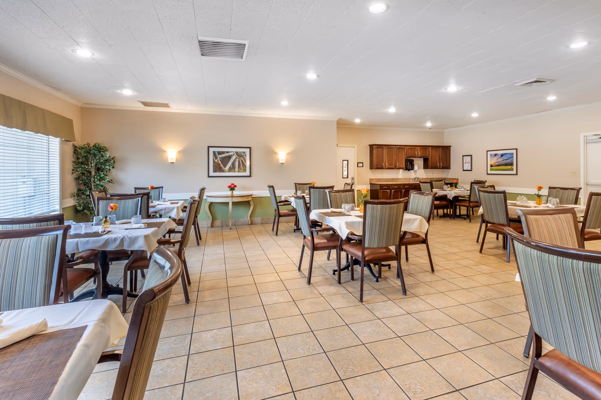 Bright tile-floored dining room with multiple set tables and chairs, a serving counter and wall art.