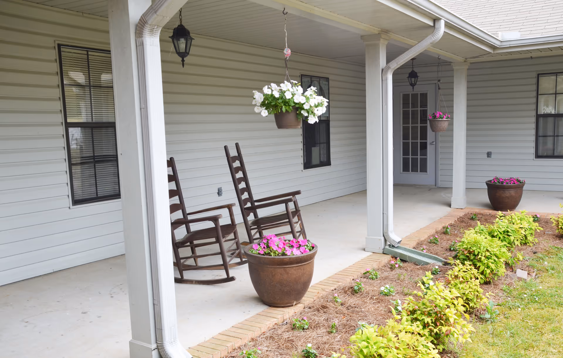 Covered porch area with two wooden rocking chairs, hanging flower pots with white and pink flowers, large flower pots on the ground, and a garden bed with green plants and small flowers along the edge of the porch. The porch is attached to a building with light gray siding and several windows and a door.
