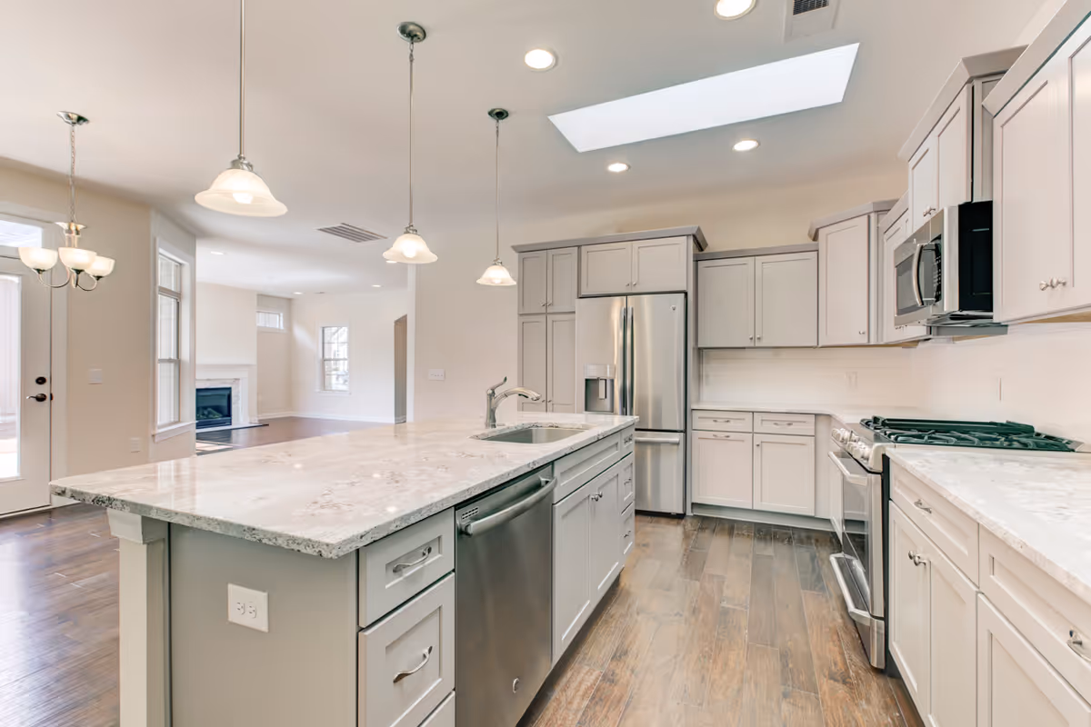 Bright modern kitchen with white cabinets, stainless steel appliances including a refrigerator, microwave, and stove. A large marble countertop island with a built-in sink and dishwasher is in the center. Pendant lights hang above the island, and a skylight provides natural light. The kitchen opens to a living area with a fireplace and windows.