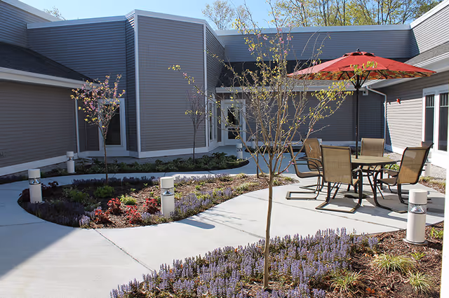 Sunny landscaped courtyard with a patio table and chairs under a red umbrella, young trees, pathways, and gray-sided building walls.