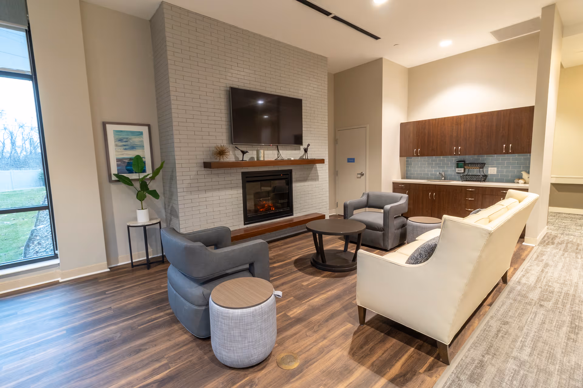 A cozy living room area with two gray armchairs, a cream-colored sofa, a round coffee table, and two small side tables. The room features a white brick fireplace with a mounted flat-screen TV above it. There is a wooden shelf above the fireplace with decorative items. To the right, there is a kitchenette area with dark wooden cabinets and a blue tiled backsplash. A large window on the left side lets in natural light, and a framed artwork and a potted plant are placed near the window.