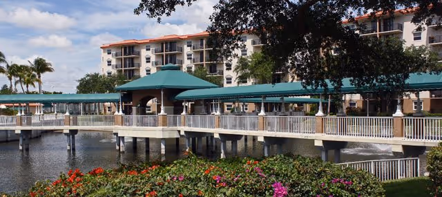 View of a senior living facility named Fountainview with a large building in the background, a covered walkway with a green roof extending over a pond, and colorful flowers and greenery in the foreground under a partly cloudy sky.
