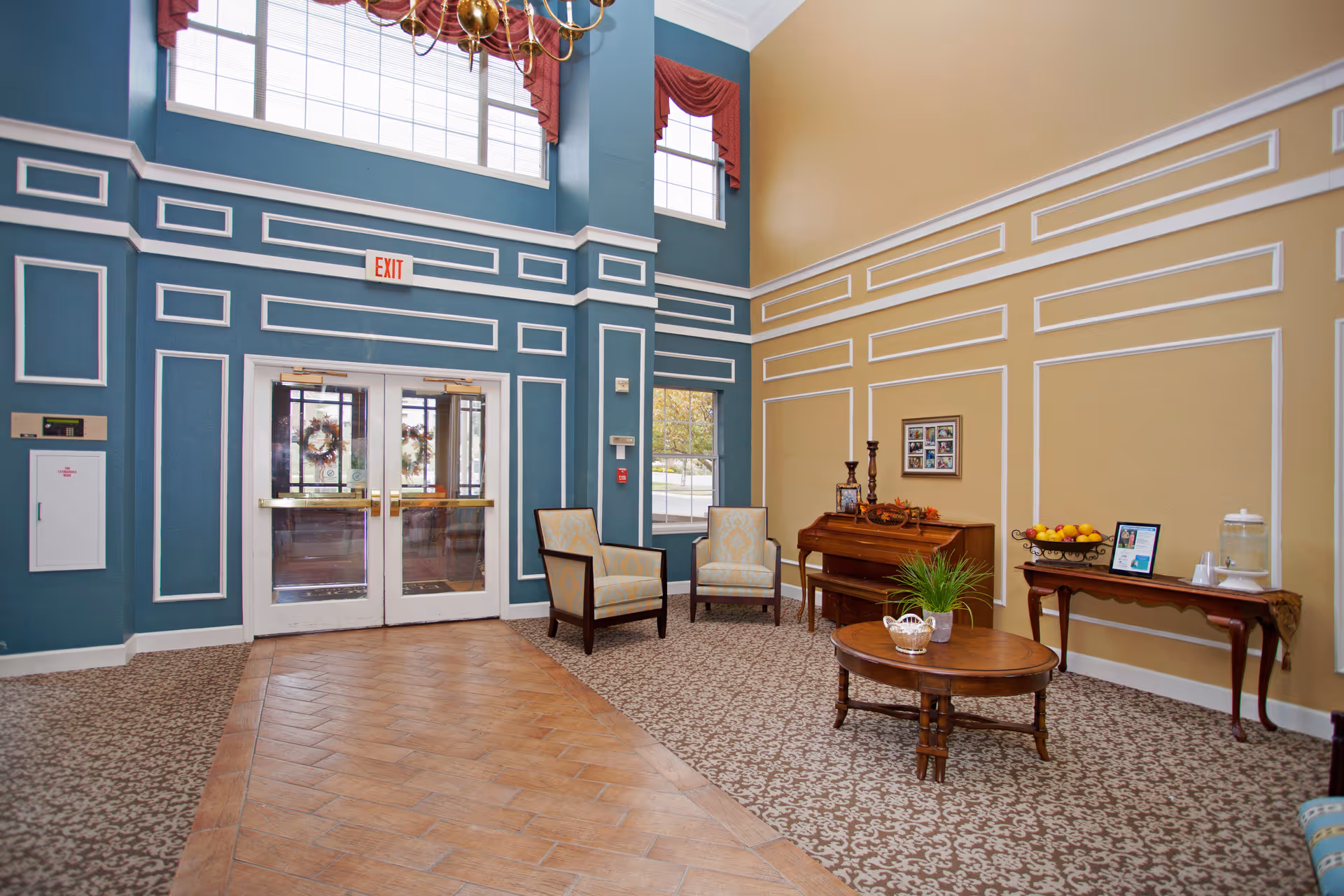 A spacious and elegant interior common area with high ceilings and large windows. The walls are painted blue and beige with white decorative molding. There are two upholstered armchairs, a wooden coffee table with a plant, a piano with decorative items, and a side table with a fruit basket and water dispenser. Double glass doors with exit signs lead outside.