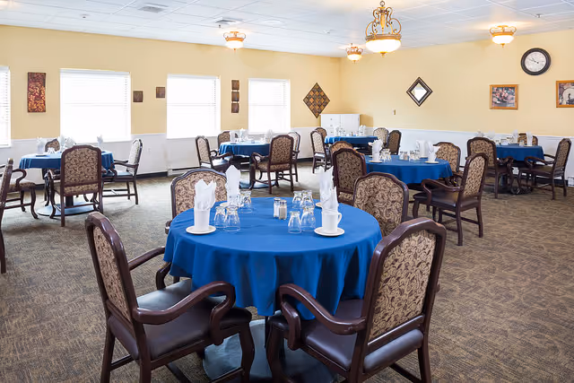 A dining room with multiple round tables covered with blue tablecloths, each set with white napkins, glasses, and cups. The room has beige walls, carpeted floors, several windows with white blinds, and decorative wall art. There are upholstered chairs with wooden frames around each table, and ceiling lights provide illumination.