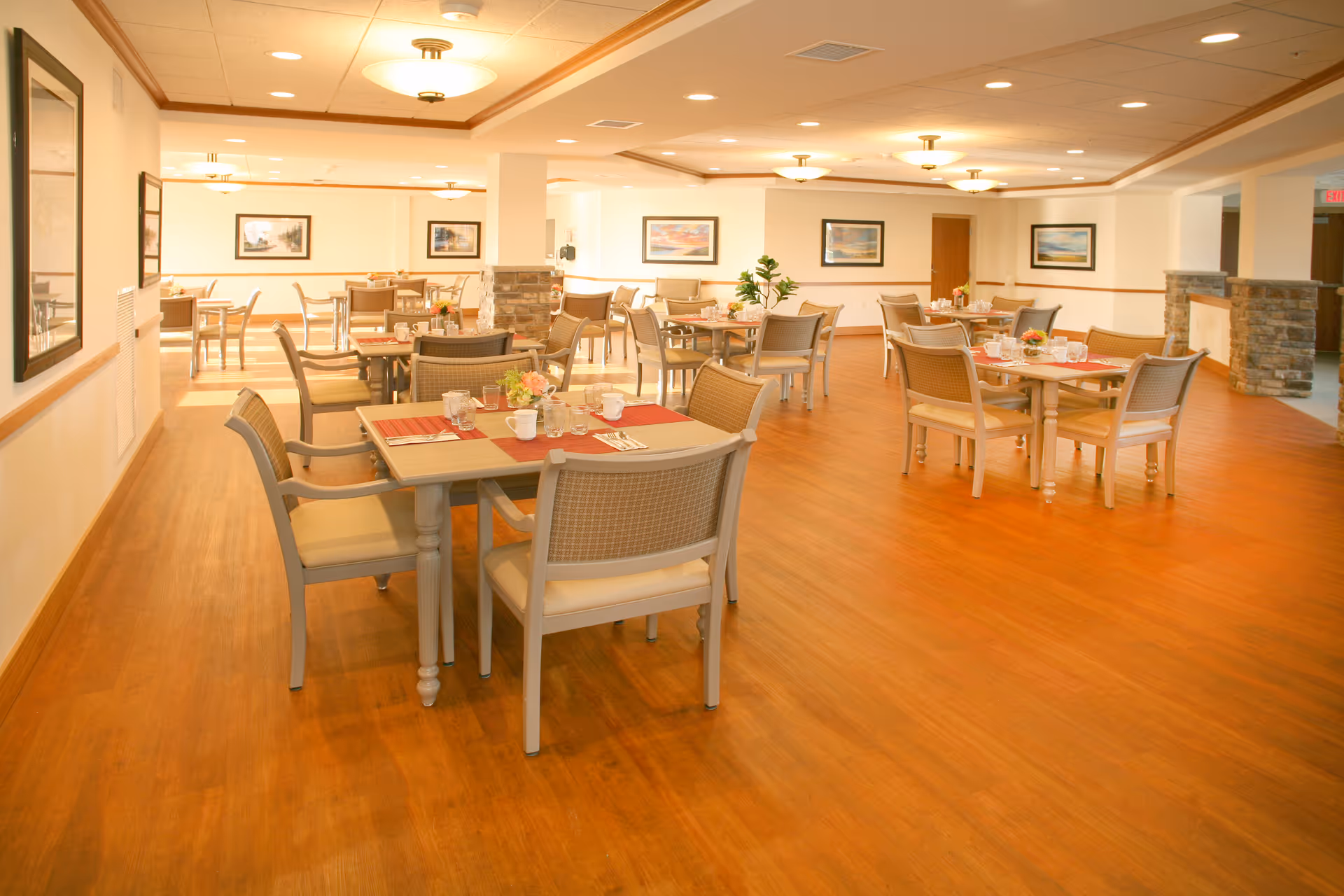 A spacious, well-lit dining room with multiple tables and chairs arranged neatly. Each table is set with placemats, cups, glasses, and small flower arrangements. The room has wooden flooring, light-colored walls, framed artwork, and ceiling lights providing warm illumination.
