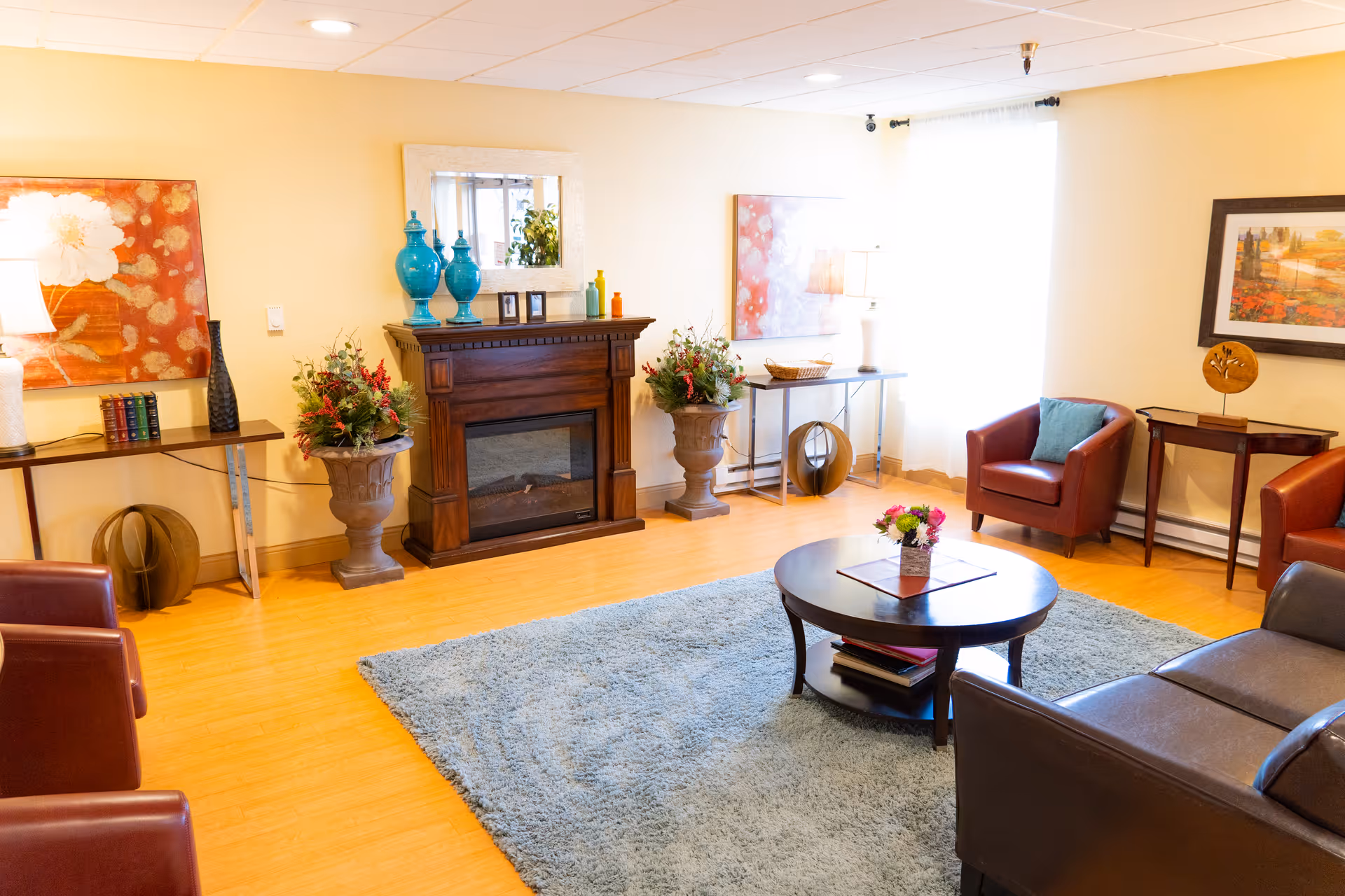 A cozy living room with a wooden floor, a light blue rug, a round wooden coffee table with a small flower arrangement, a dark brown leather sofa, and two red leather armchairs. The room features a wooden electric fireplace with decorative vases and a mirror above it. There are two side tables with lamps and decorative items, and framed artwork on the walls. Natural light comes through a window with sheer white curtains.
