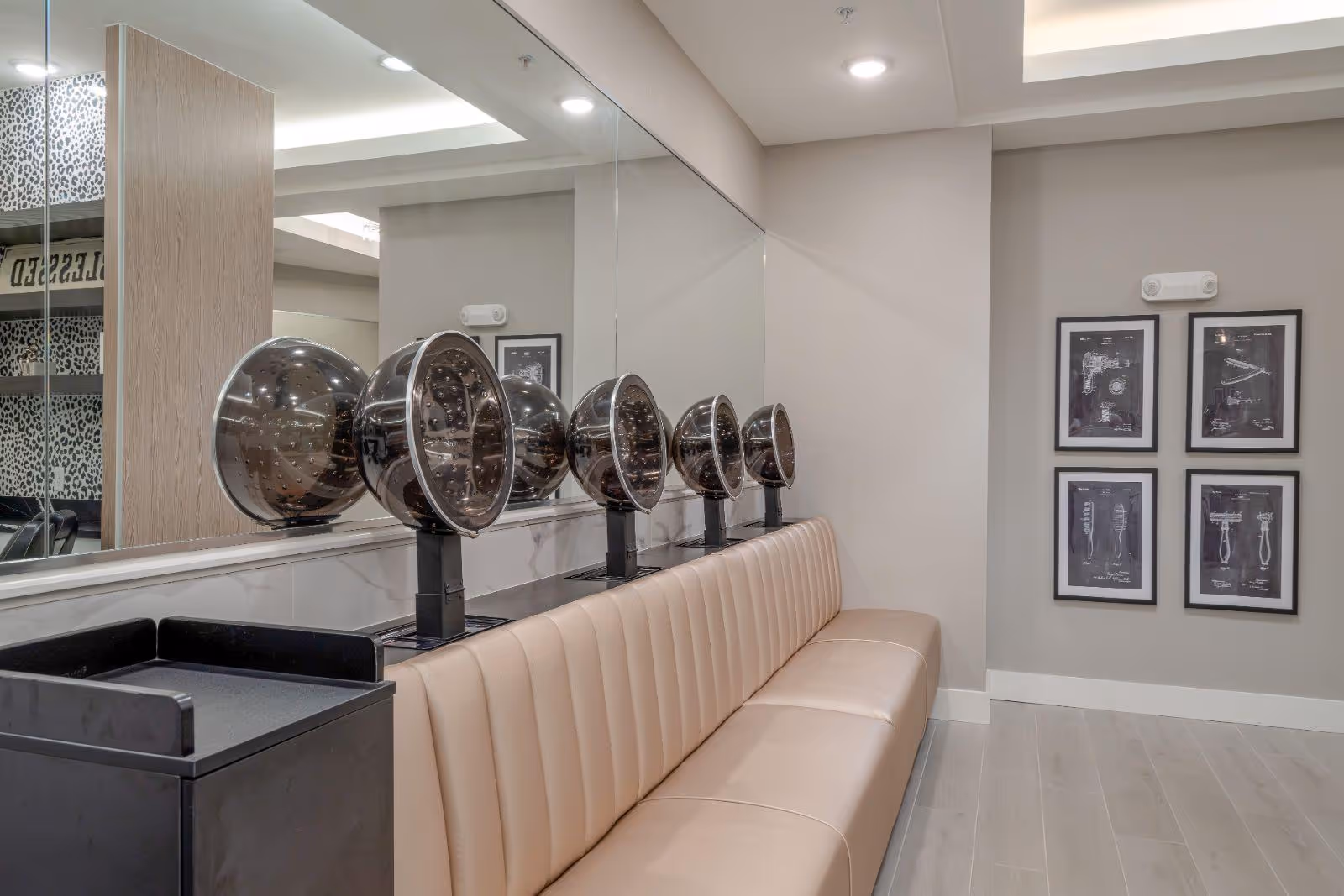 Interior salon area with padded bench seating, multiple hooded hair dryers mounted along a mirrored wall, and framed artwork on the opposite wall.