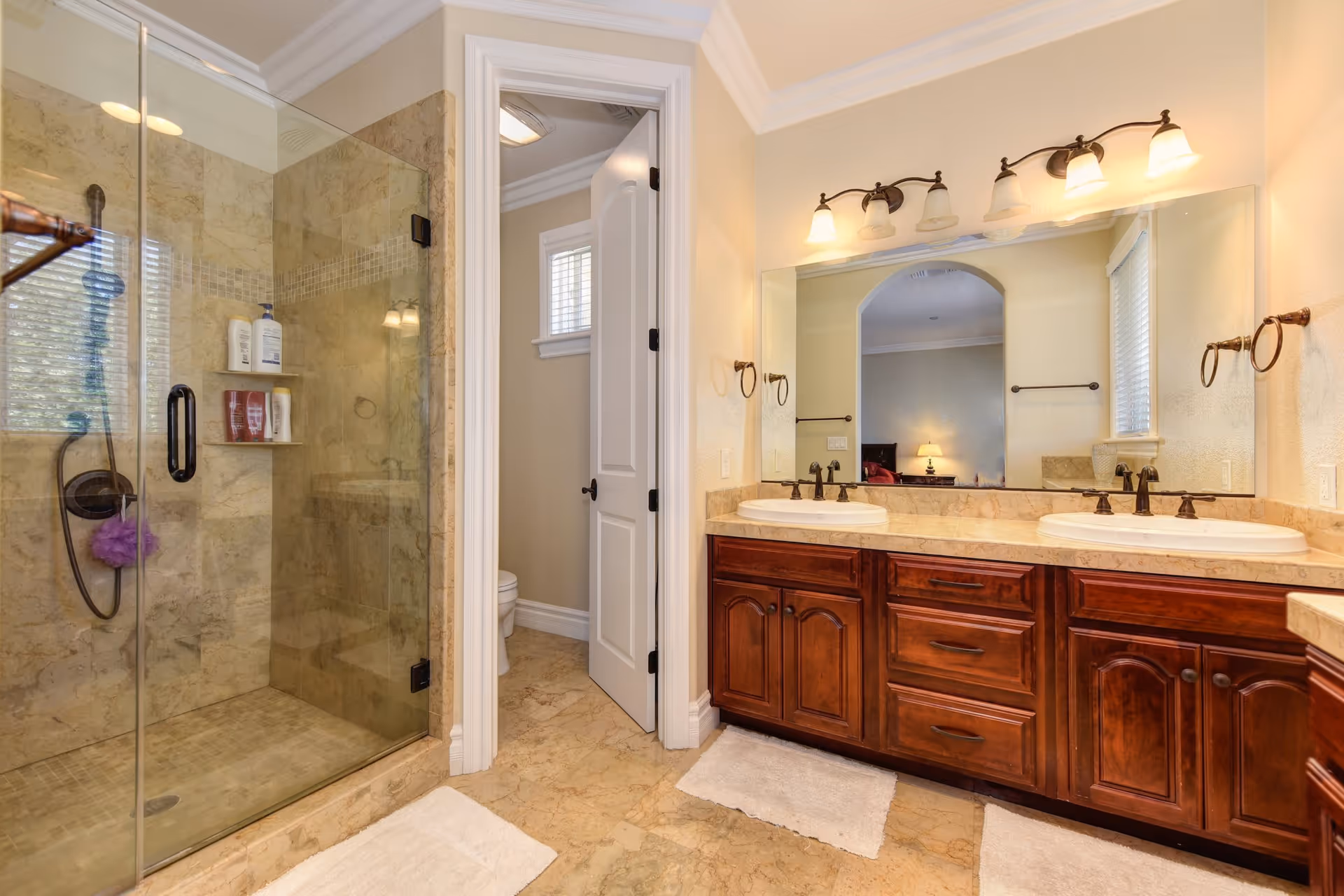 A spacious bathroom featuring a glass-enclosed shower with beige marble tiles, a double sink vanity with dark wood cabinets, and a large mirror above the sinks. The bathroom has light-colored walls and floor tiles, with white bath mats on the floor. A door next to the vanity leads to a separate toilet area with a small window.