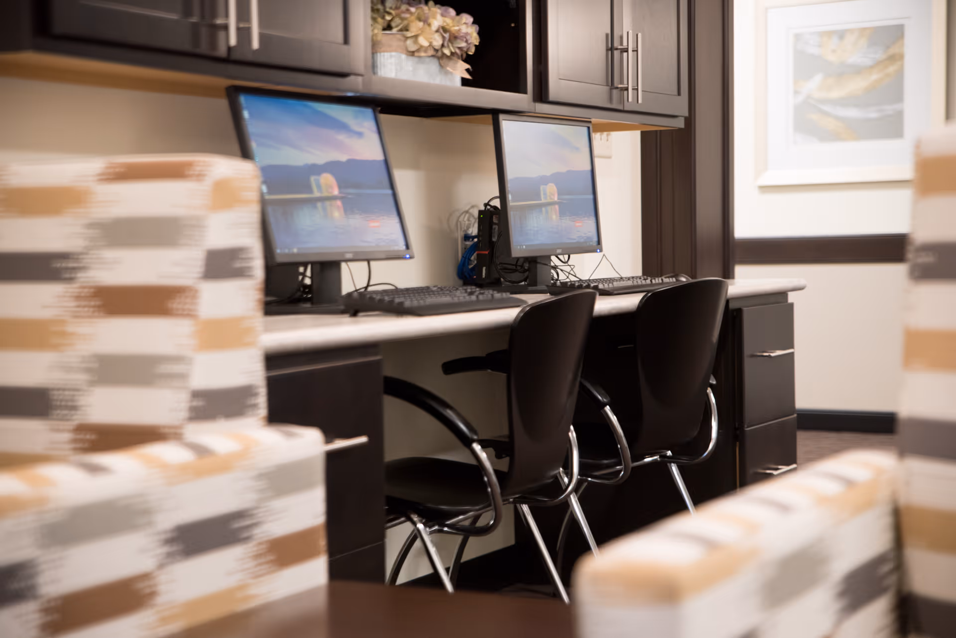 A small computer workstation area with two desktop computers and black chairs, set against a wall with dark wood cabinets above. In the foreground, there are patterned upholstered chairs in shades of beige, brown, and gray. A framed picture hangs on the wall in the background.