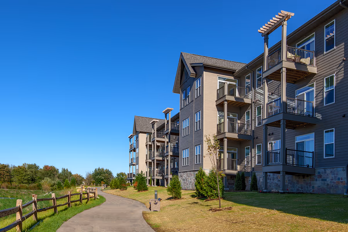 Exterior view of a multi-story residential building with balconies, adjacent to a paved walking path bordered by a wooden fence and green grass under a clear blue sky.