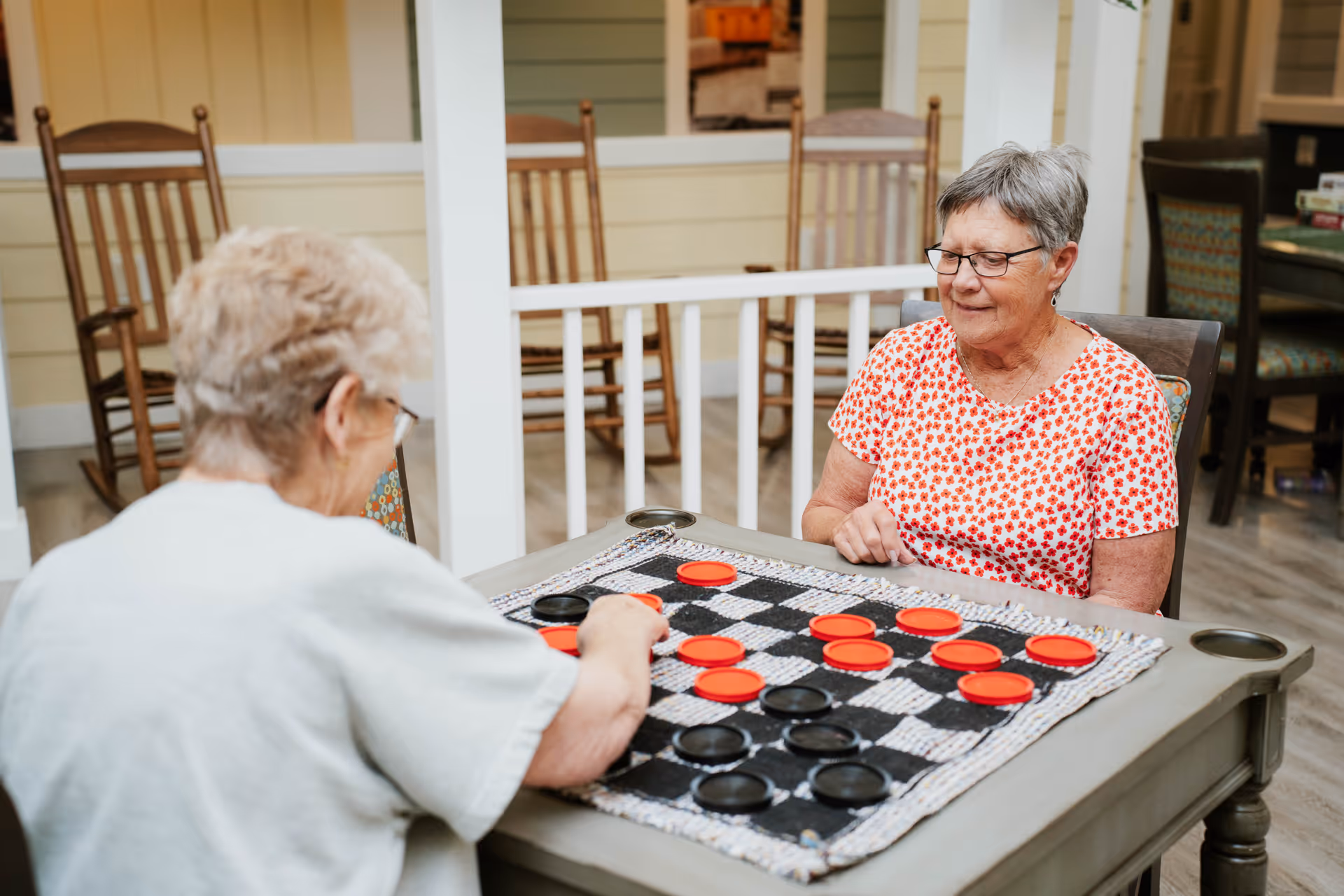 Two elderly women sitting at a table playing a game of checkers indoors. One woman is wearing a white shirt and glasses, seen from behind, while the other woman, wearing a white shirt with red floral patterns and glasses, is facing the camera and smiling. The room has wooden rocking chairs and a light-colored interior.