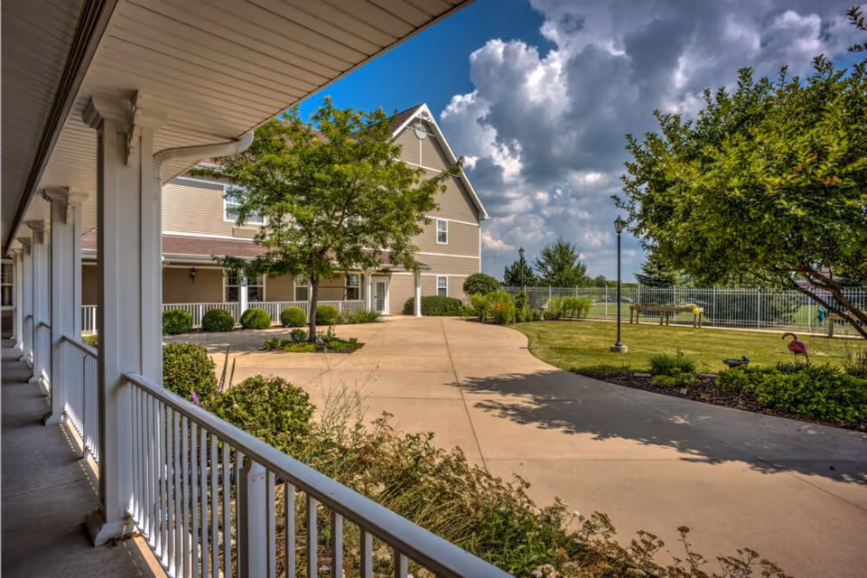 View of a senior living facility exterior walkway with white railings and columns on the left, leading to a beige building with multiple windows. There is a paved pathway surrounded by landscaped greenery, trees, and bushes under a partly cloudy sky.