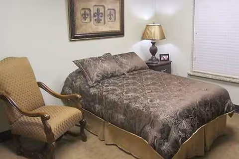 A cozy bedroom in a senior living facility with a patterned bed, chair, nightstand, and lamp.