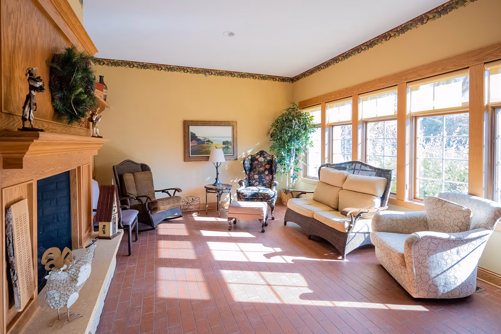 A bright living room with large windows letting in sunlight. The room features a wooden fireplace decorated with a wreath and small sculptures, several chairs including a floral armchair, a cushioned loveseat, and a patterned armchair. There is a small table with a lamp and a potted plant near the windows. The floor is made of reddish-brown tiles and the walls are painted a light yellow with a decorative border near the ceiling.