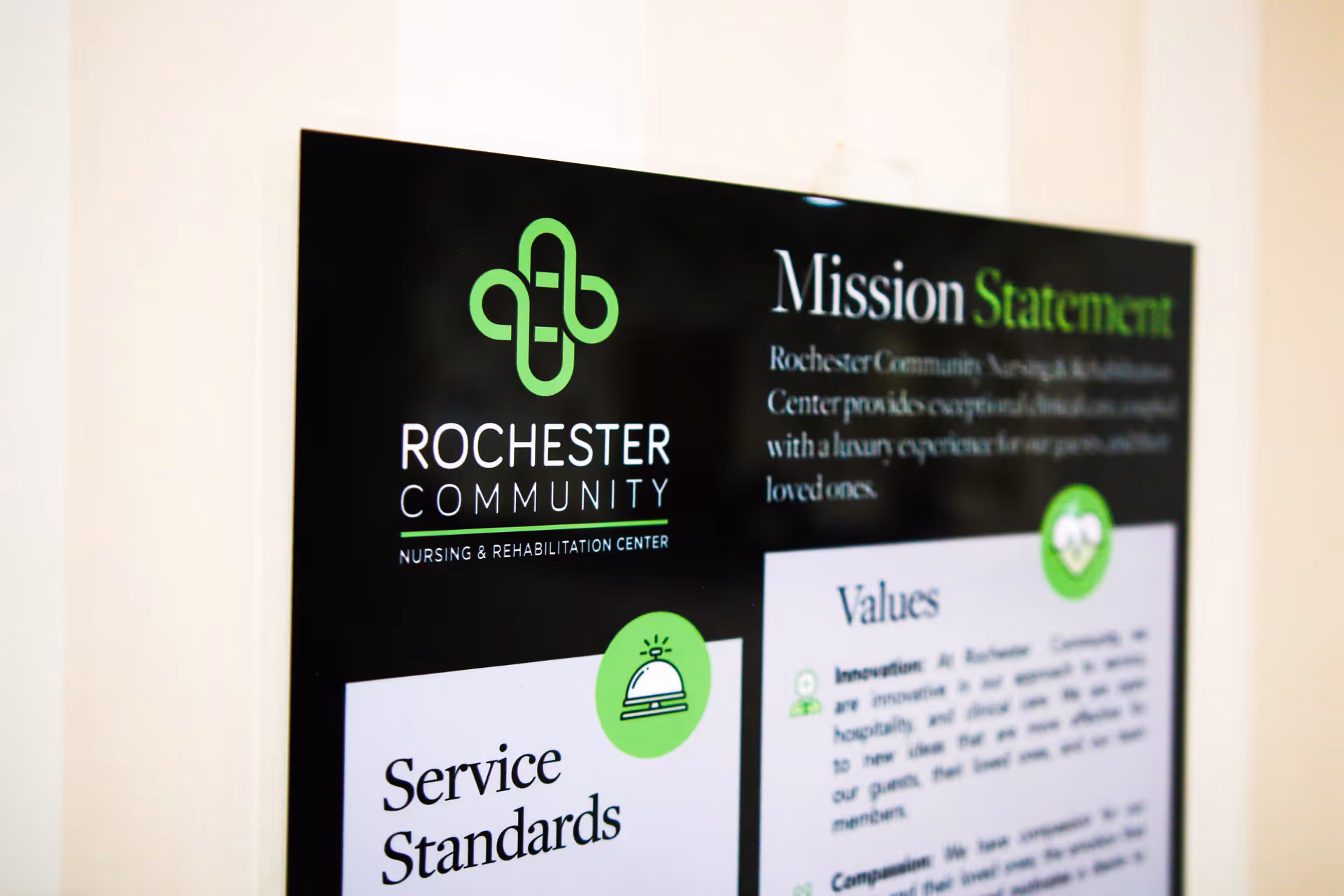 A close-up view of a wall-mounted informational sign for Rochester Community Nursing & Rehabilitation Center displaying their mission statement, service standards, and values with a black and green design.