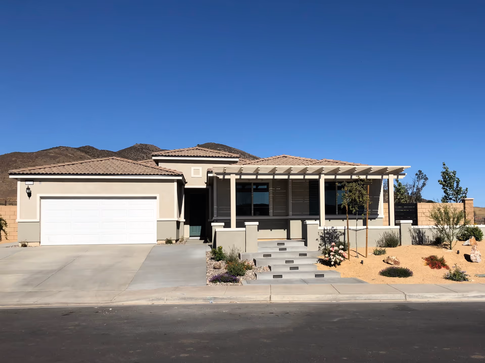 Single-story modern house with a tiled roof, a two-car garage, a front porch with a pergola, and desert landscaping with small plants and rocks. Mountains are visible in the background under a clear blue sky.