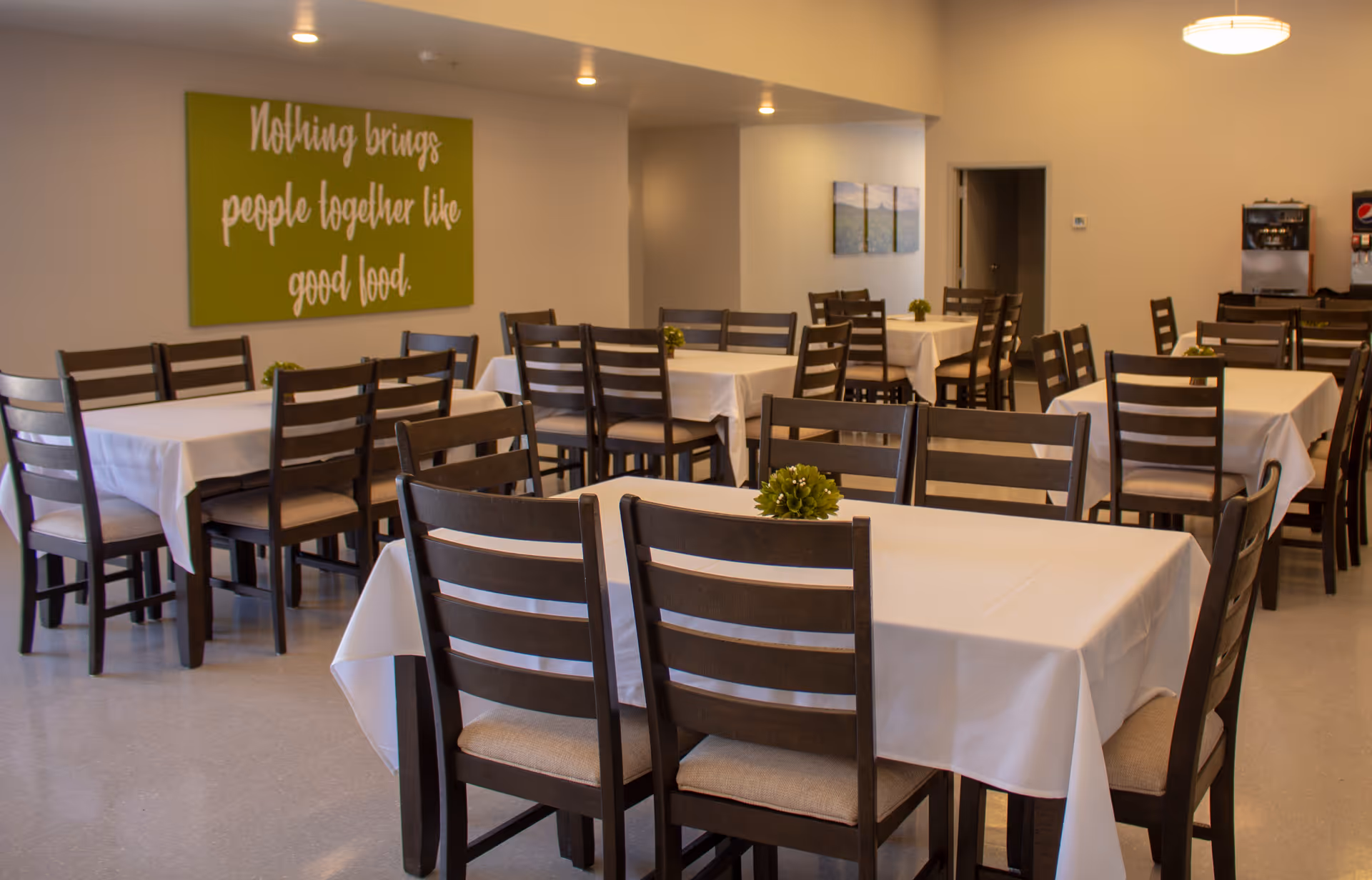 A dining room with multiple tables covered in white tablecloths and surrounded by wooden chairs with cushioned seats. Each table has a small green plant centerpiece. On the wall, there is a green sign with white text that reads, 'Nothing brings people together like good food.' In the background, there is a beverage dispenser and some wall art.