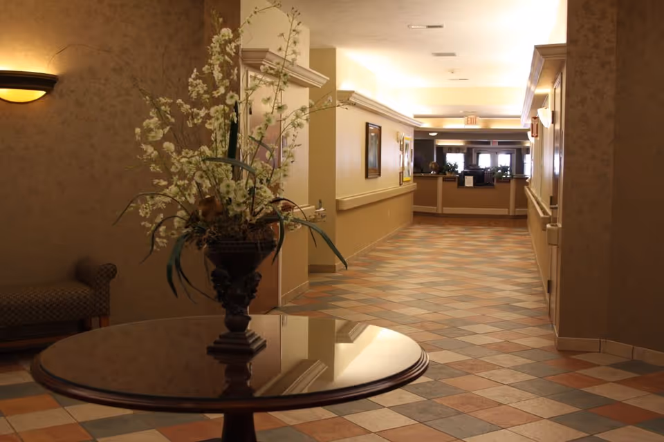 A well-lit hallway in a senior living facility with patterned tile flooring and beige walls. A round wooden table with a decorative floral arrangement is in the foreground on the left side. The hallway leads to a reception desk area with plants and framed artwork on the walls.