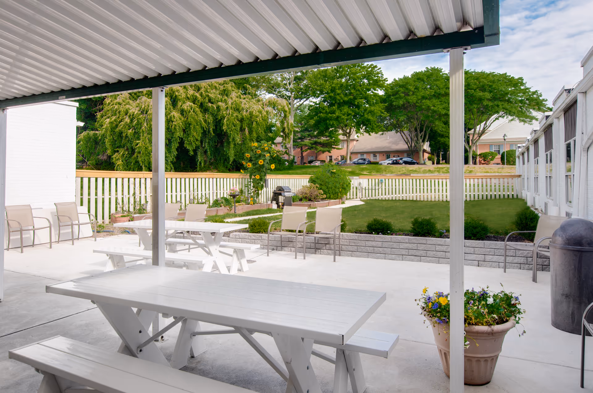 Covered outdoor patio area with white picnic tables and benches, beige chairs along the sides, a flower pot with colorful flowers, and a green lawn with trees and a white picket fence in the background.