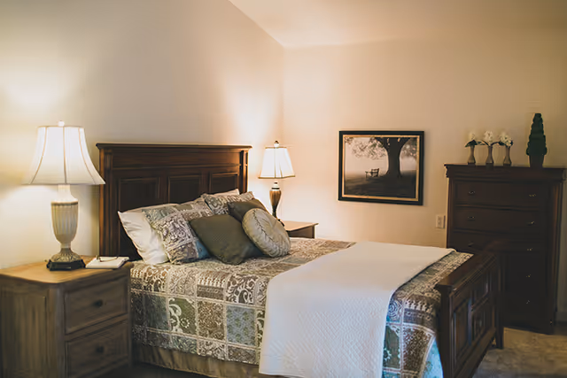 Cozy bedroom with a wooden bed, patterned bedspread, nightstands with lamps, a dresser, and framed artwork on the wall.