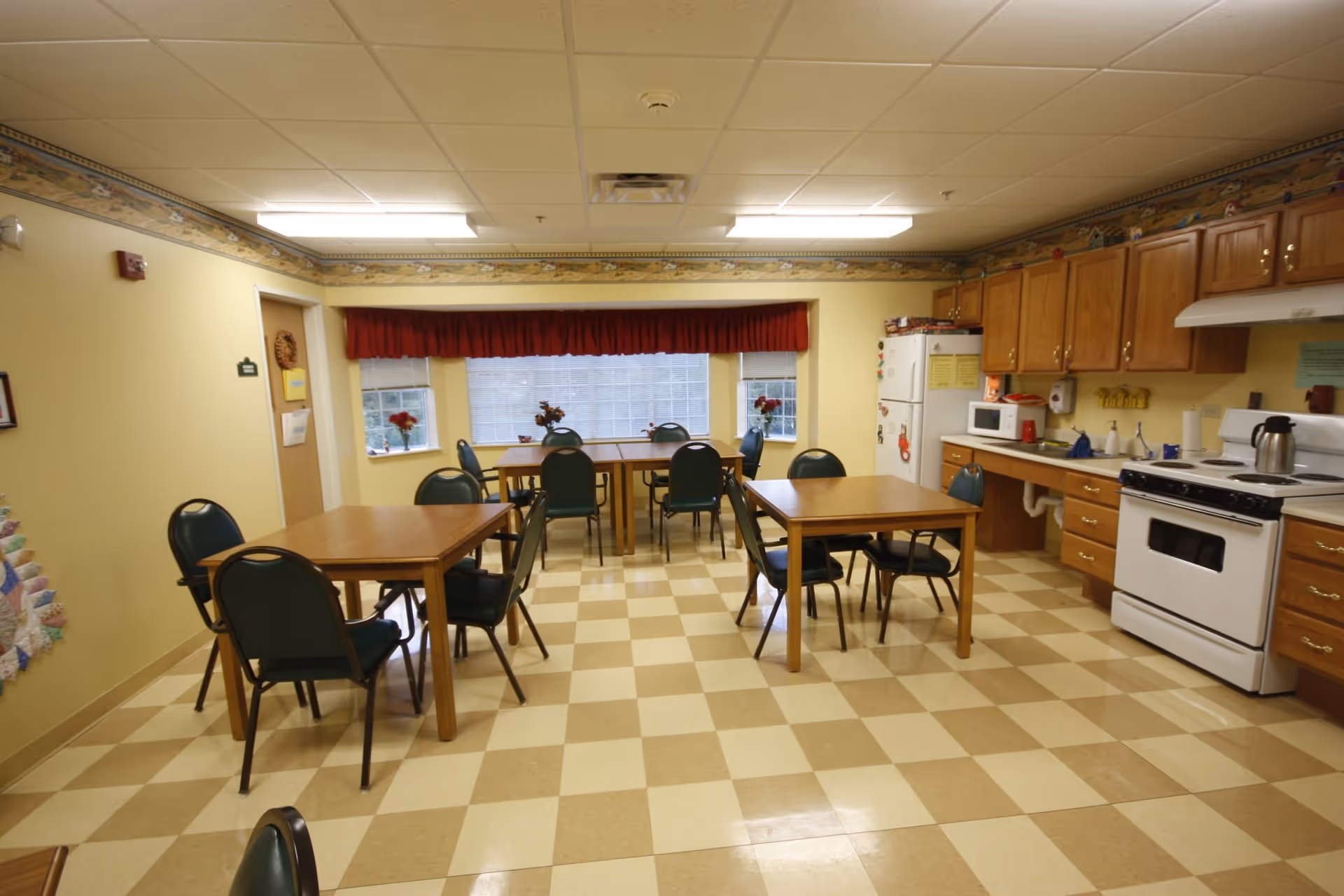 Communal dining room with several tables and chairs and a kitchenette with stove and refrigerator.